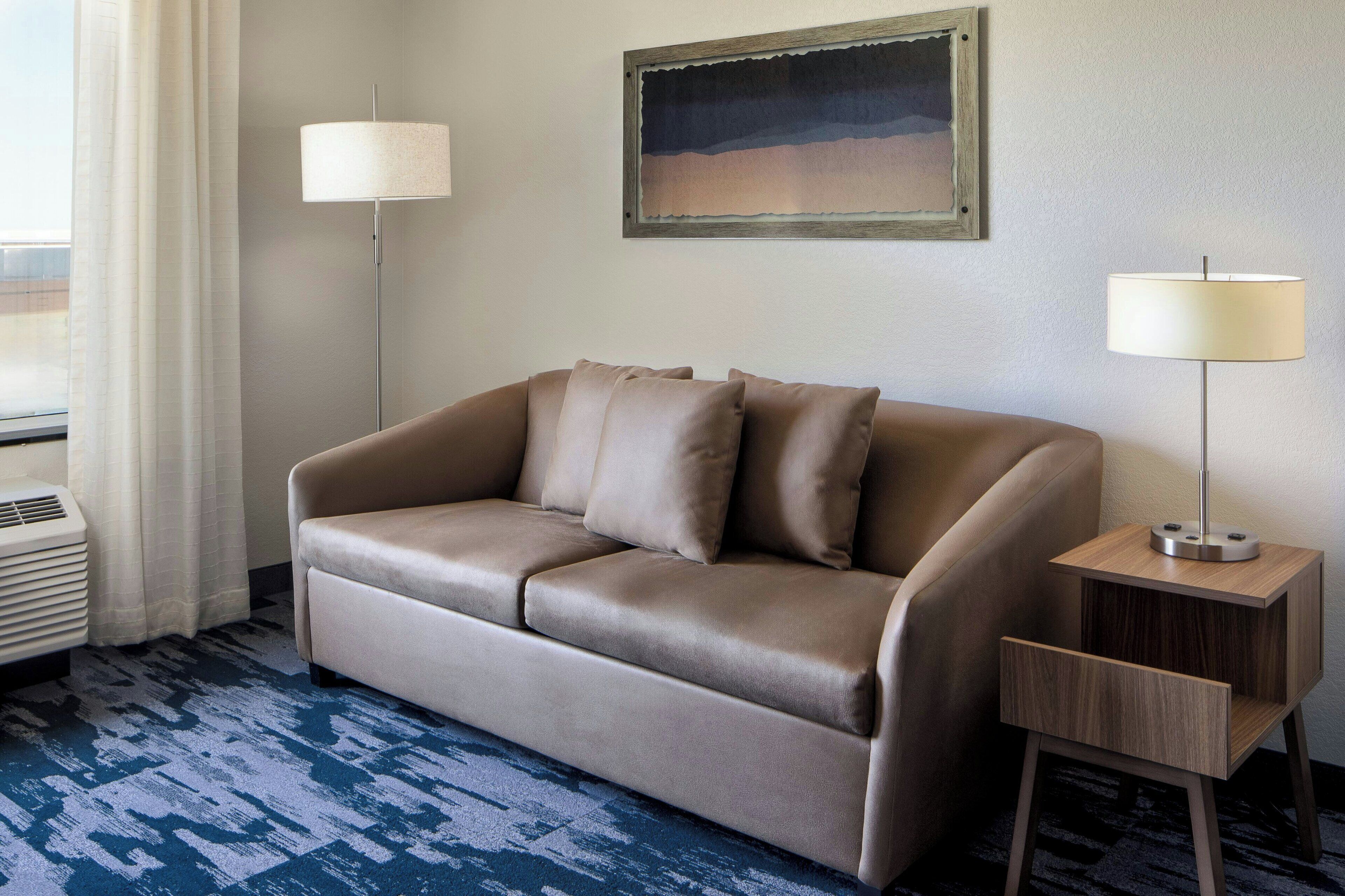 A modern hotel room with a beige leather sofa, a side table with a lamp, a framed picture on the wall, and a blue patterned carpet.