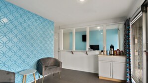 A modern room with a blue patterned wall, a grey armchair, a small round table, a white cabinet with kitchen items, and a window with a view.