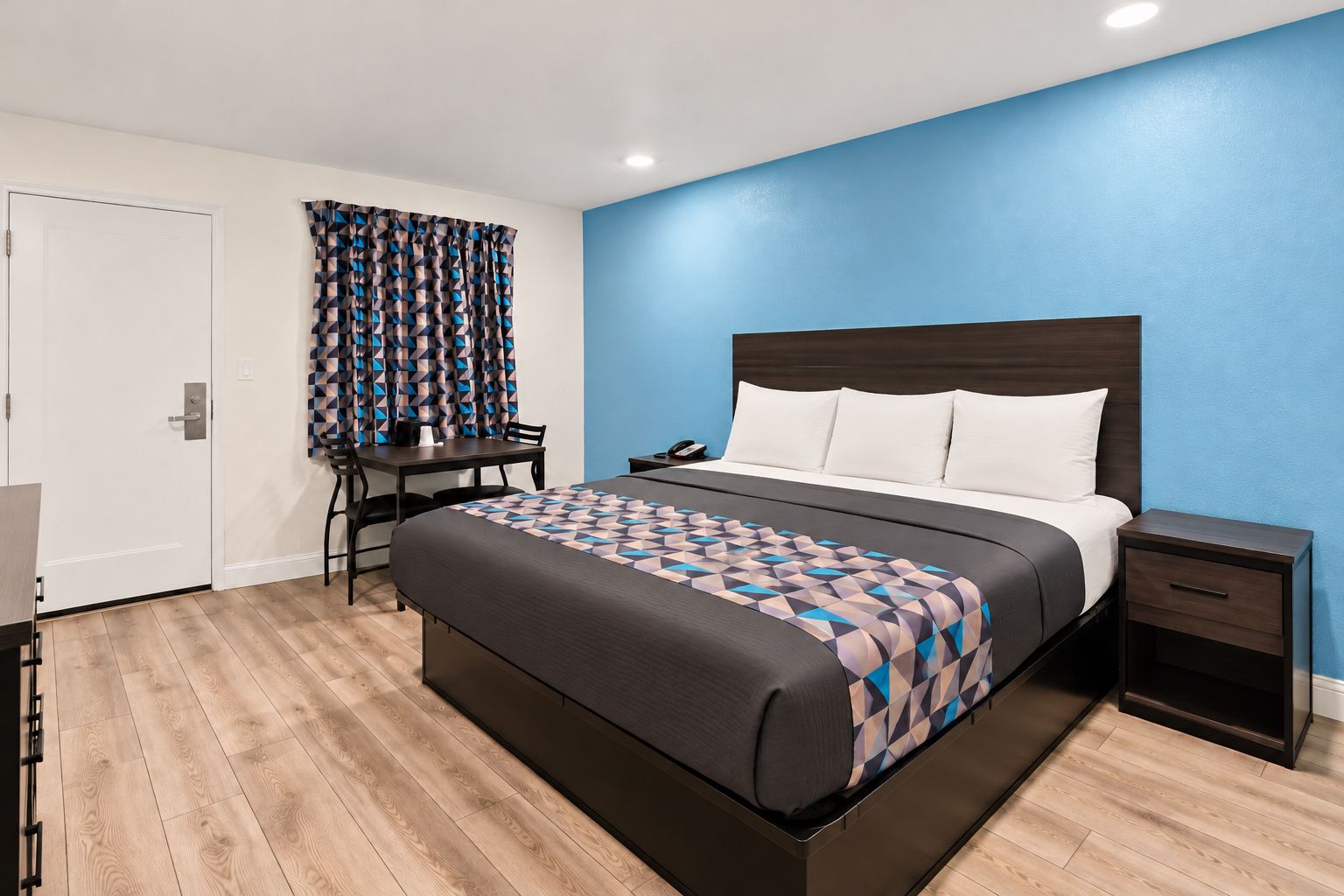 A hotel room with a bed, bedside table, desk, chair, and a patterned bedspread.