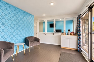 Una habitación moderna con pared azul estampada, una mesita redonda pequeña, dos sillas, una kitchenette con armarios y una ventana con cortinas.
