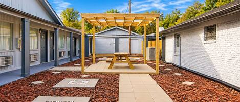 A covered patio area with a wooden pergola, surrounded by a garden with red mulch and stepping stones, and adjacent to a building with a white exterior.