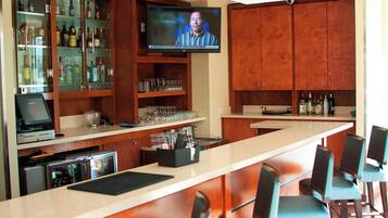 Un espace bar doté d’un long comptoir, de tabourets de bar, d’un téléviseur fixé au-dessus du bar, ainsi que d’étagères où sont disposées des bouteilles et des verres.