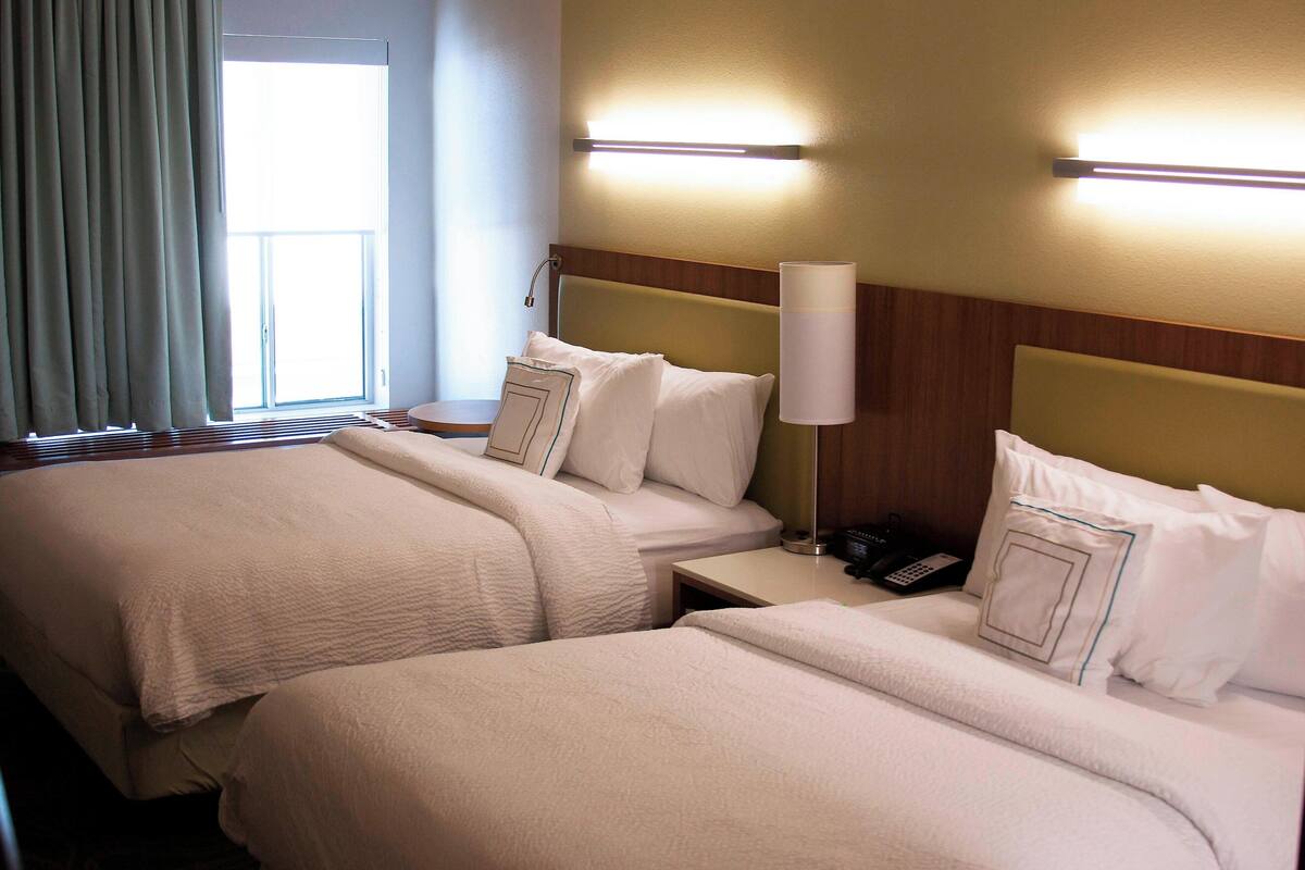 A hotel room with two beds, a bedside table with a lamp, a phone, and a window with curtains.