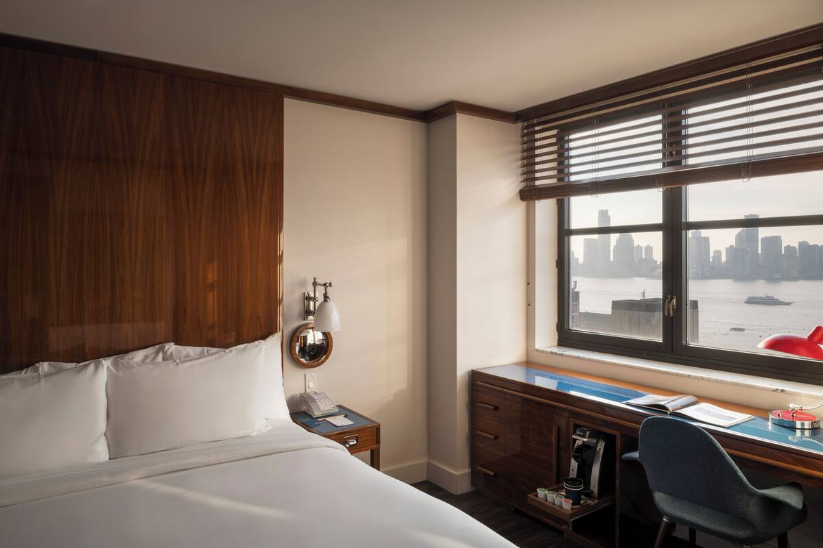 A hotel room with a bed, a desk, a chair, a window with a city view, and a wooden wardrobe.