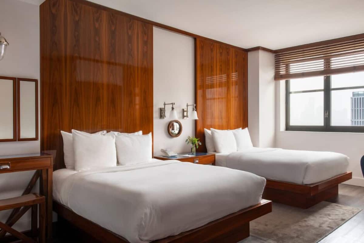 A hotel room with two beds, wooden headboards, a desk, and a large window with blinds.
