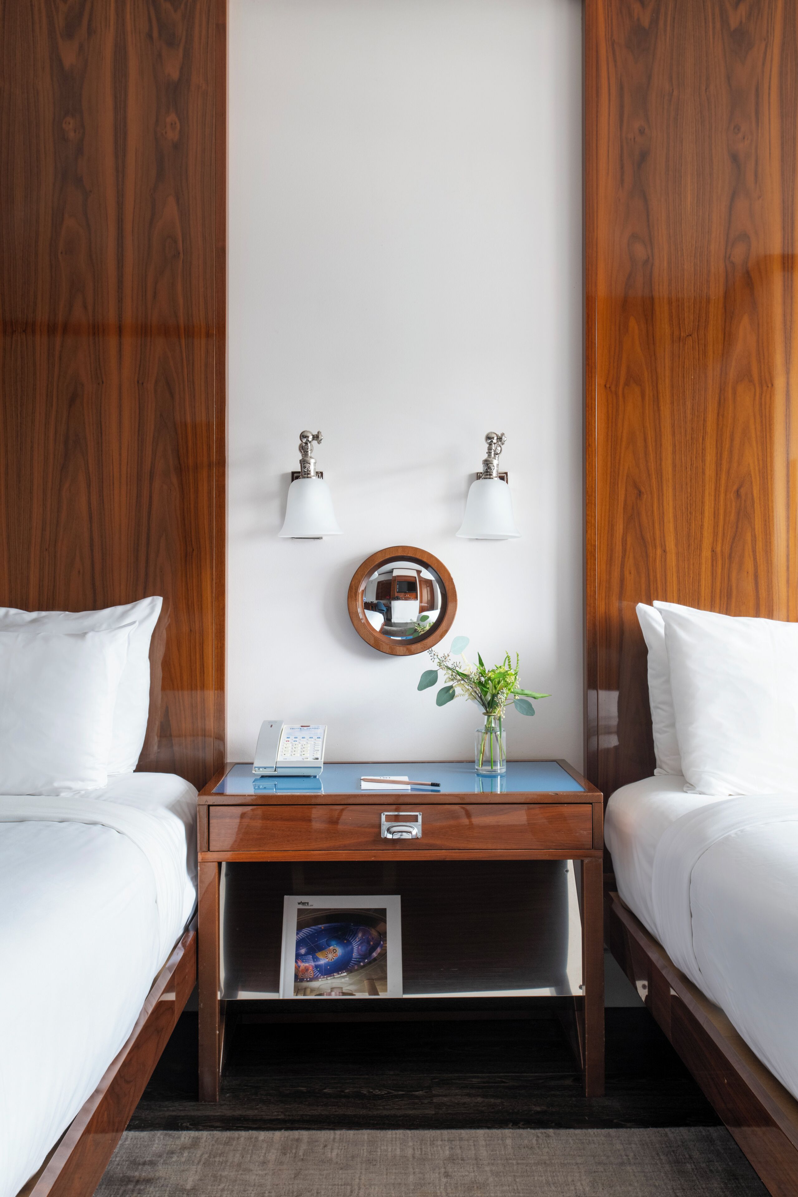 Habitación de hotel con dos camas, una mesita de noche de madera con lámpara, un espejo y una planta pequeña.