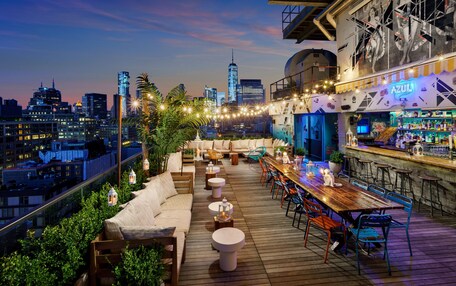 A rooftop bar with a city skyline view, wooden deck, outdoor seating, and a bar area with stools.