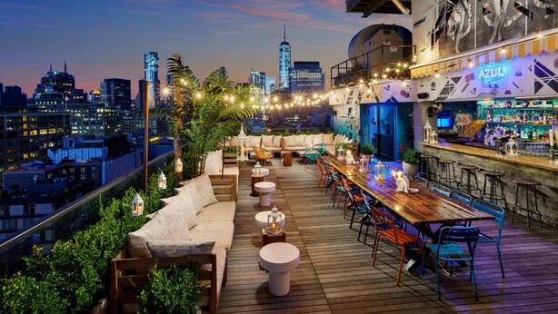 Un bar situé sur le toit, offrant une vue panoramique sur la ville, avec une terrasse en bois, des places assises en plein air et un espace bar doté de tabourets.