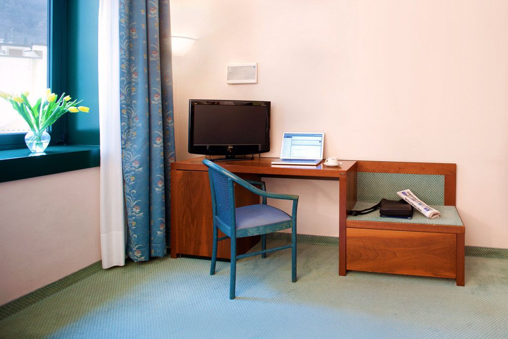 Ein Zimmer mit Schreibtisch, Stuhl, Fernseher, Laptop, einem kleinen Tisch und einer Vase mit Blumen.