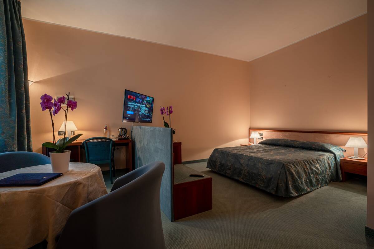 A hotel room with a bed, a desk with a computer, a chair, a small table with a plant, and a TV on the wall.
