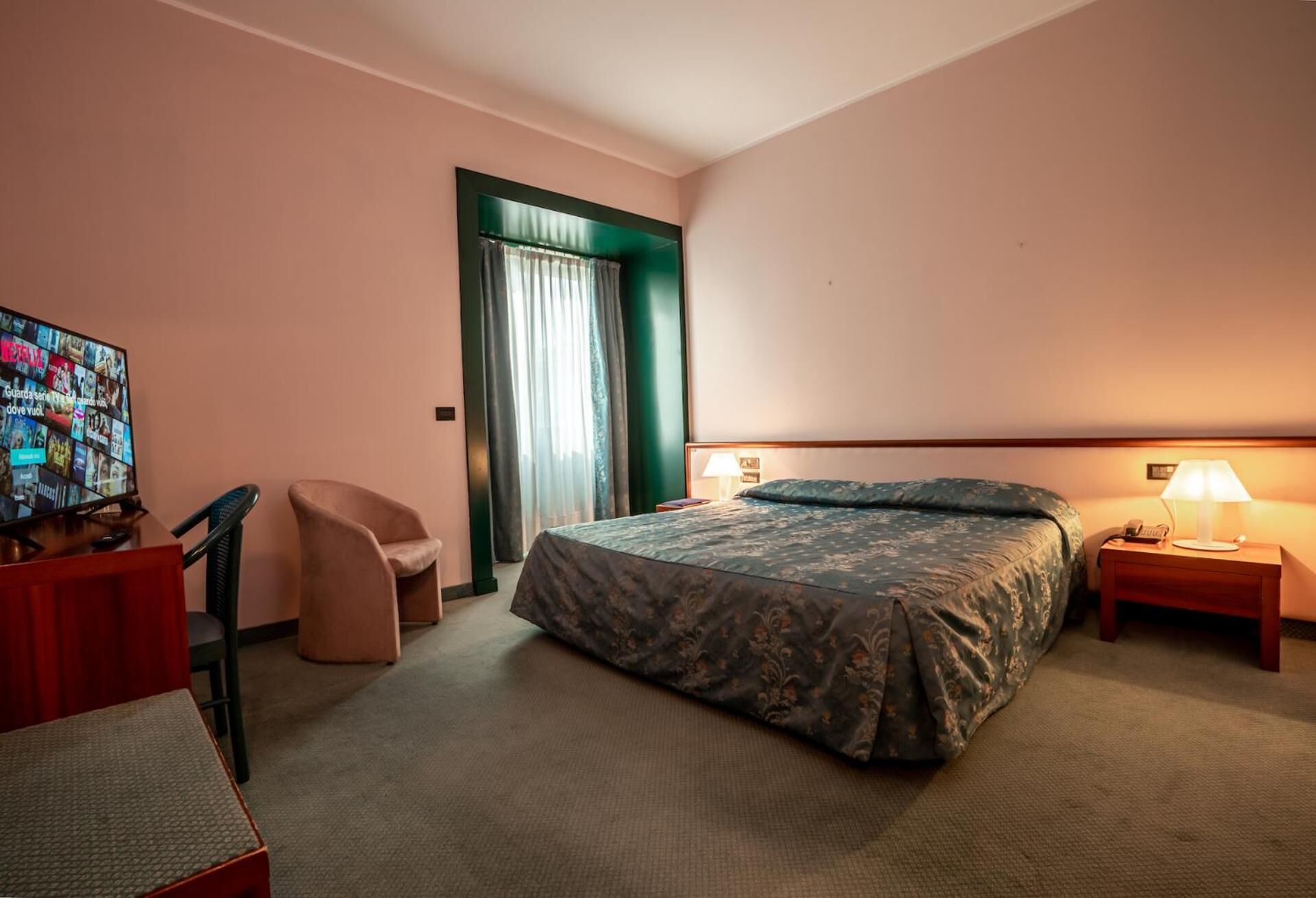 Habitación de hotel con cama, escritorio con televisor, silla y mesita de noche con lámpara.
