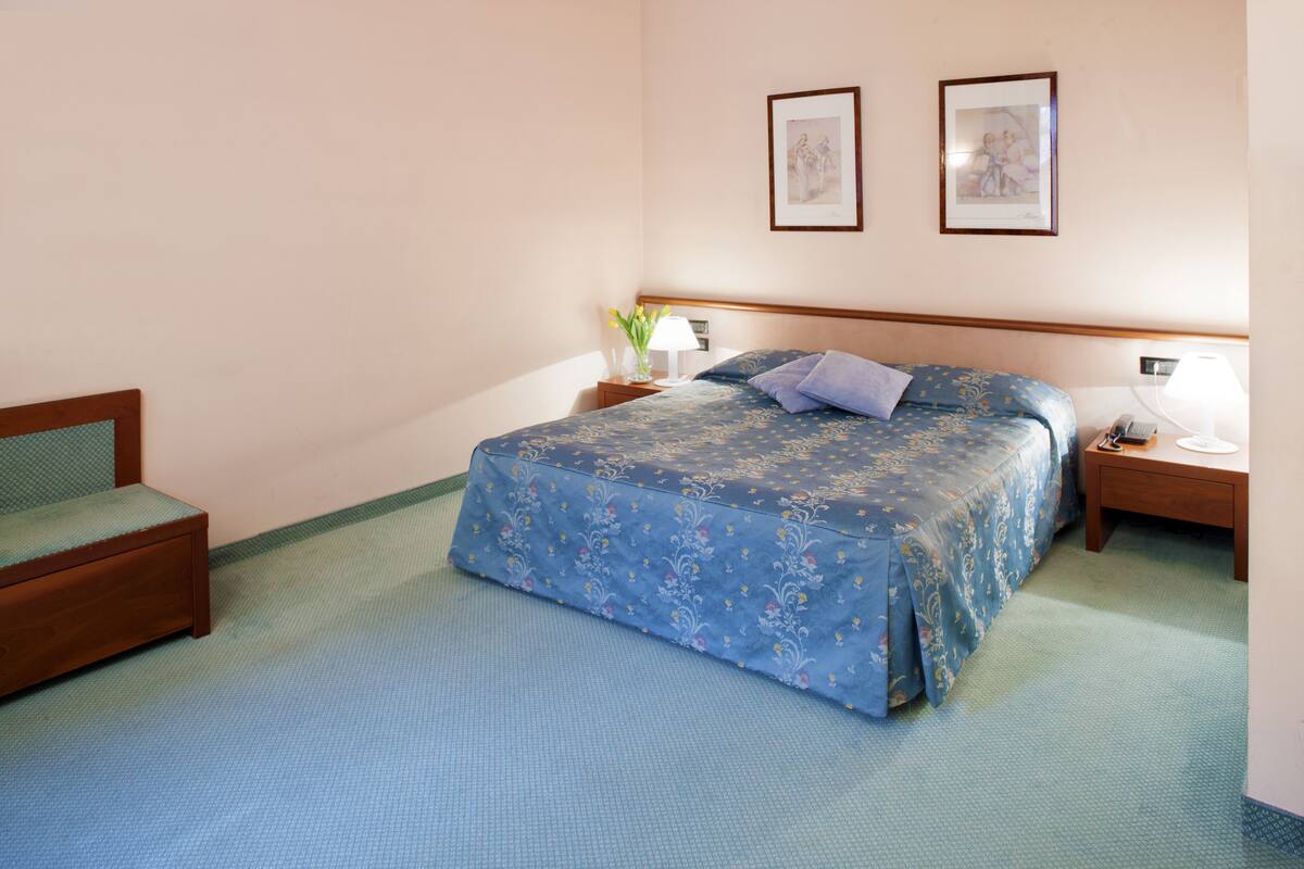 A hotel room with a bed, a chair, a nightstand, and two framed pictures on the wall.