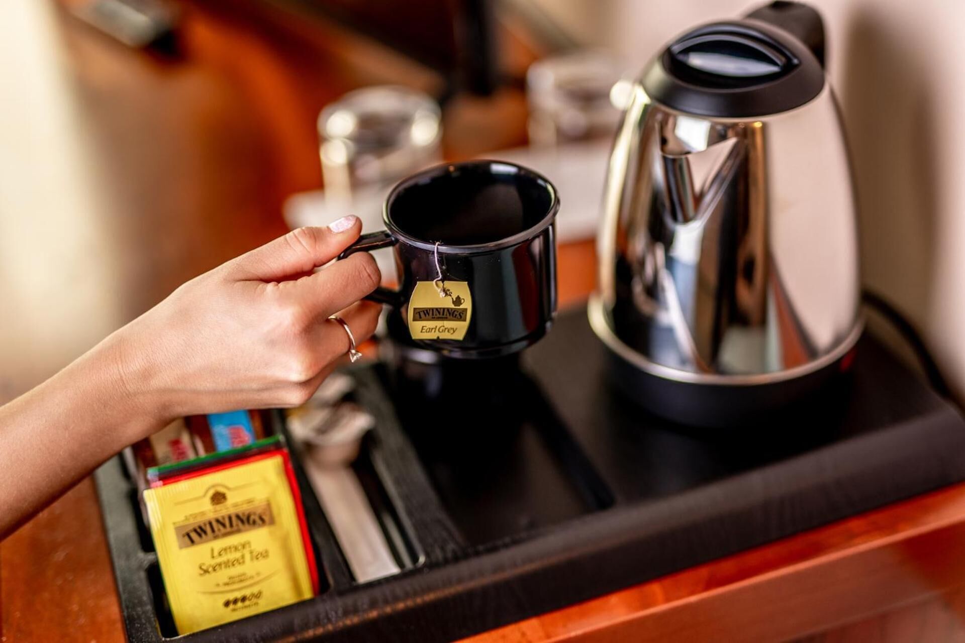 A hand holding a black mug with Earl Grey tea, a black teapot, and a box of lemon scented tea on a tray.