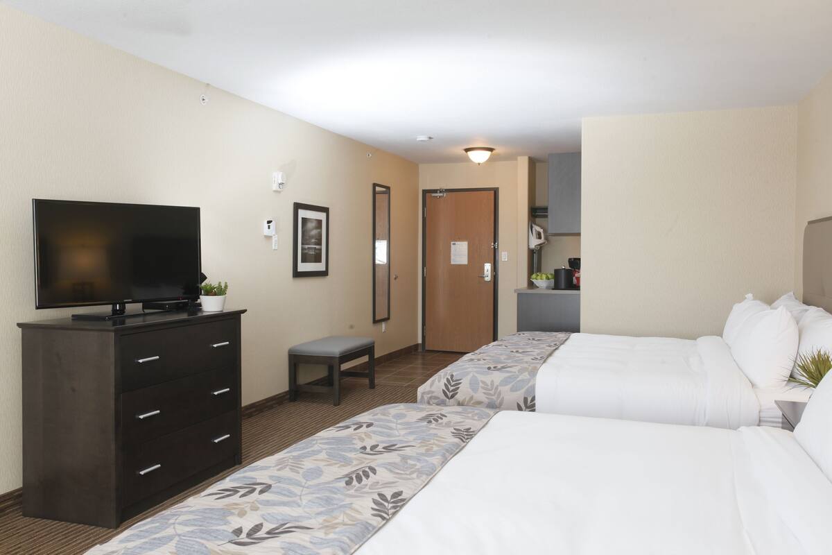 A hotel room with two beds, a TV on a dresser, a small bench, and a door leading to a kitchenette.