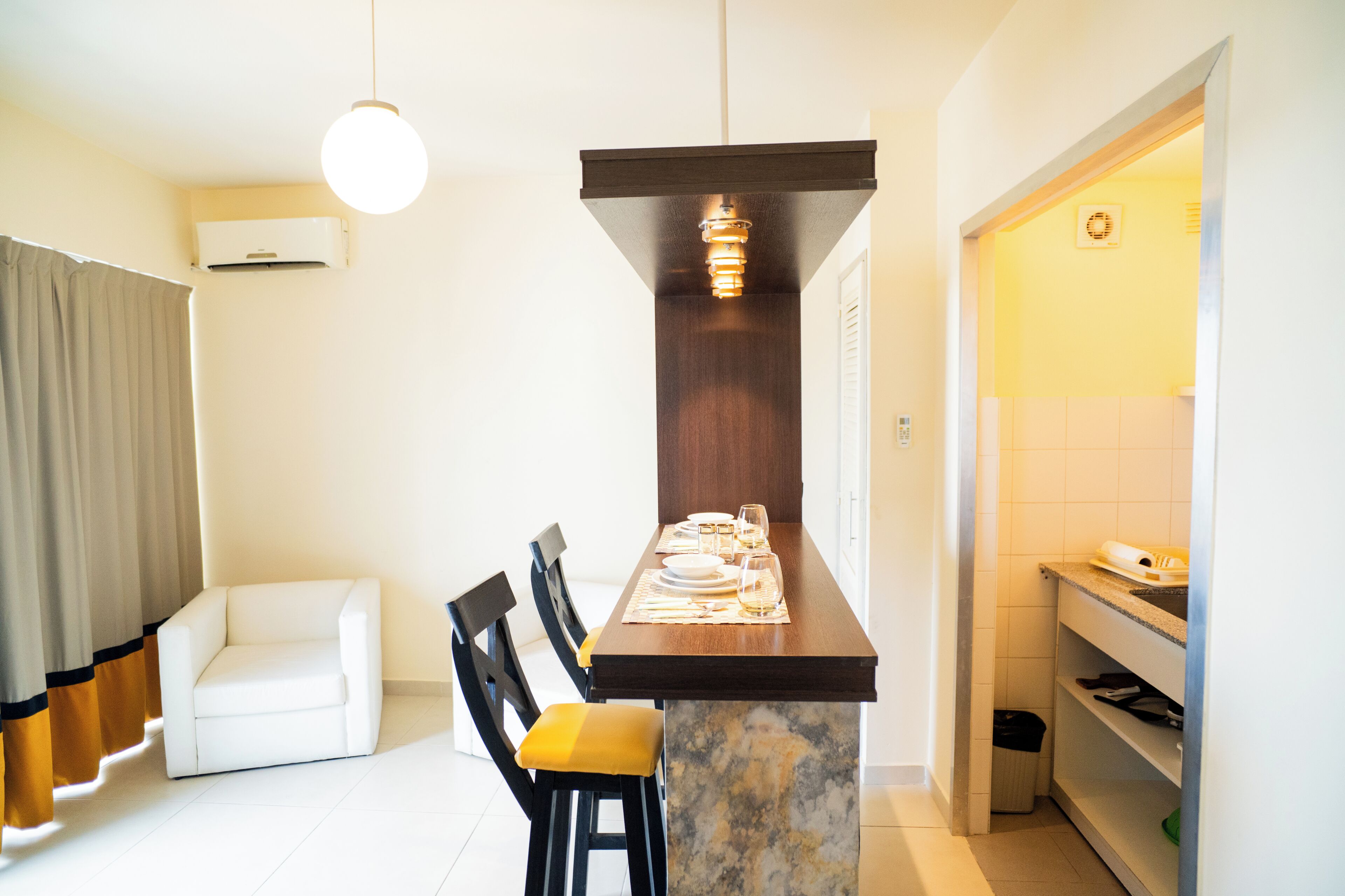 A compact kitchen with a marble countertop, a dining table set with plates and glasses, a hanging light fixture, and a white air conditioner on the wall.