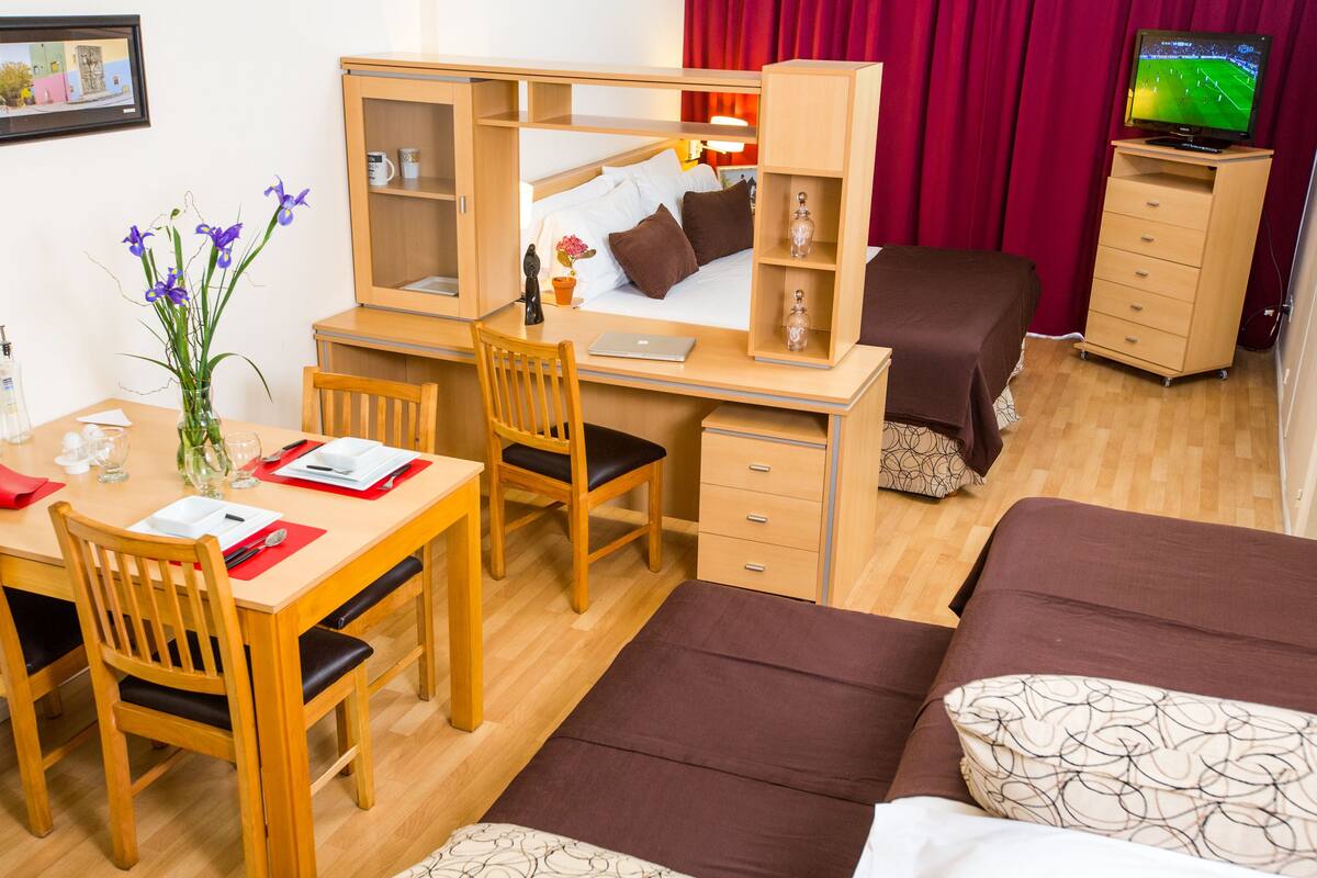 A compact hotel room with a bed, a desk, a chair, a TV, and a small dining table.