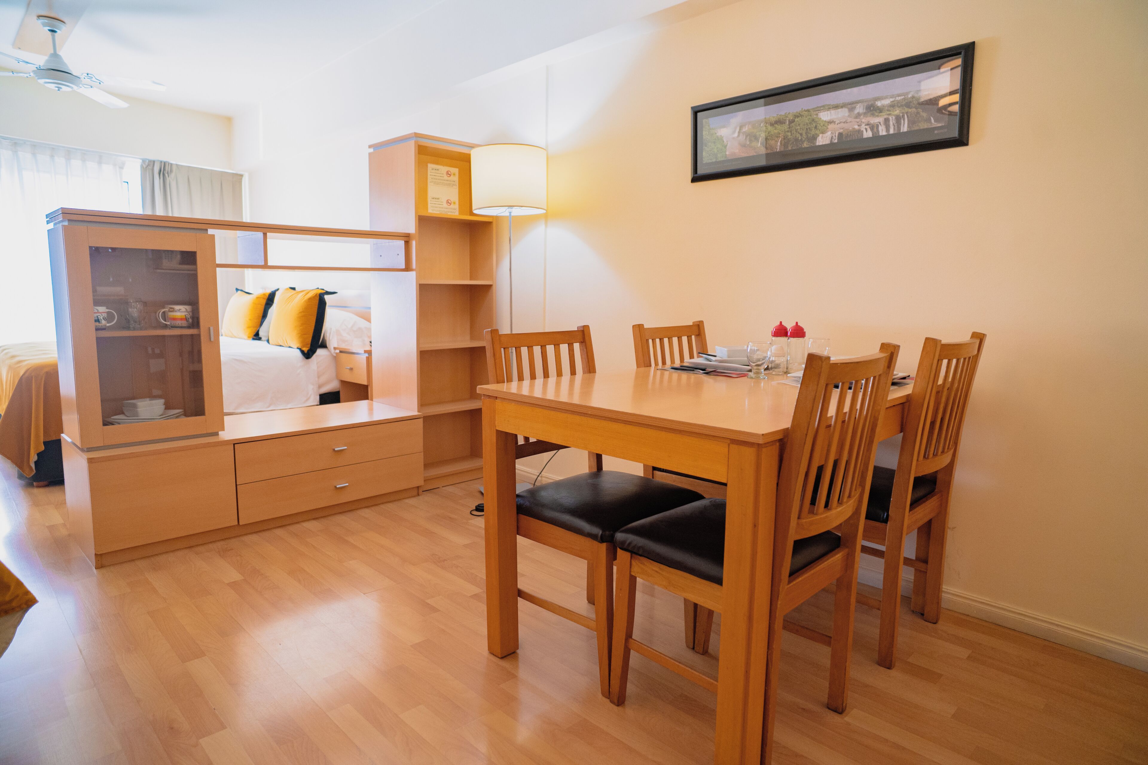 A hotel room with a wooden dining table, four chairs, a wardrobe, a bed with a white headboard, and a framed picture on the wall.