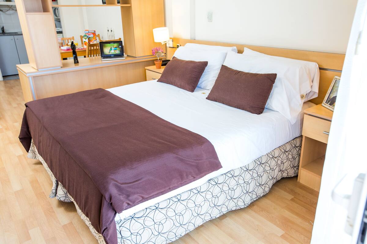 A neatly made bed with a brown bedspread and white linens, a bedside table with a lamp, and a small potted plant.
