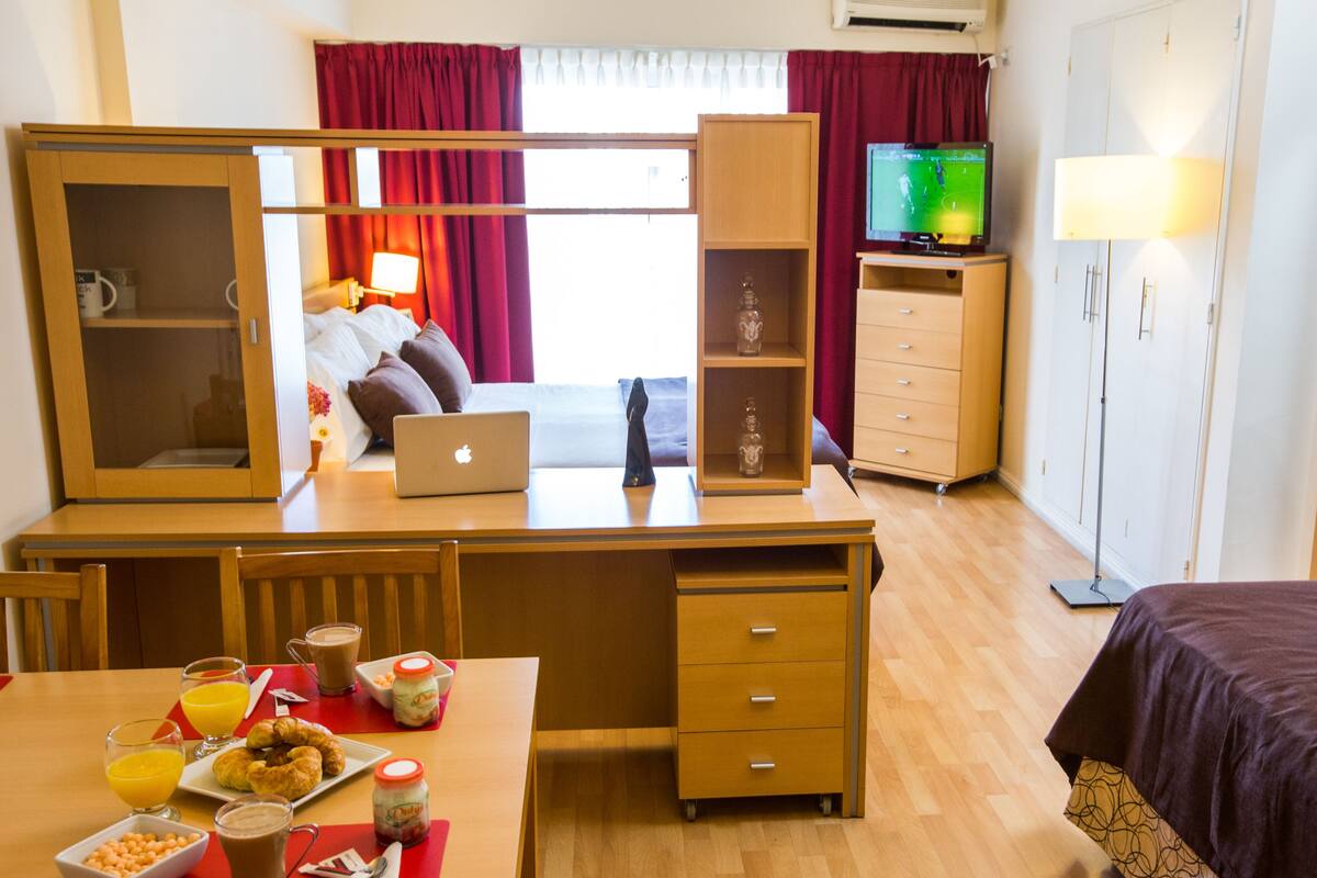 A hotel room with a wooden desk, a laptop, a television, a bed, and a dining area with breakfast items.