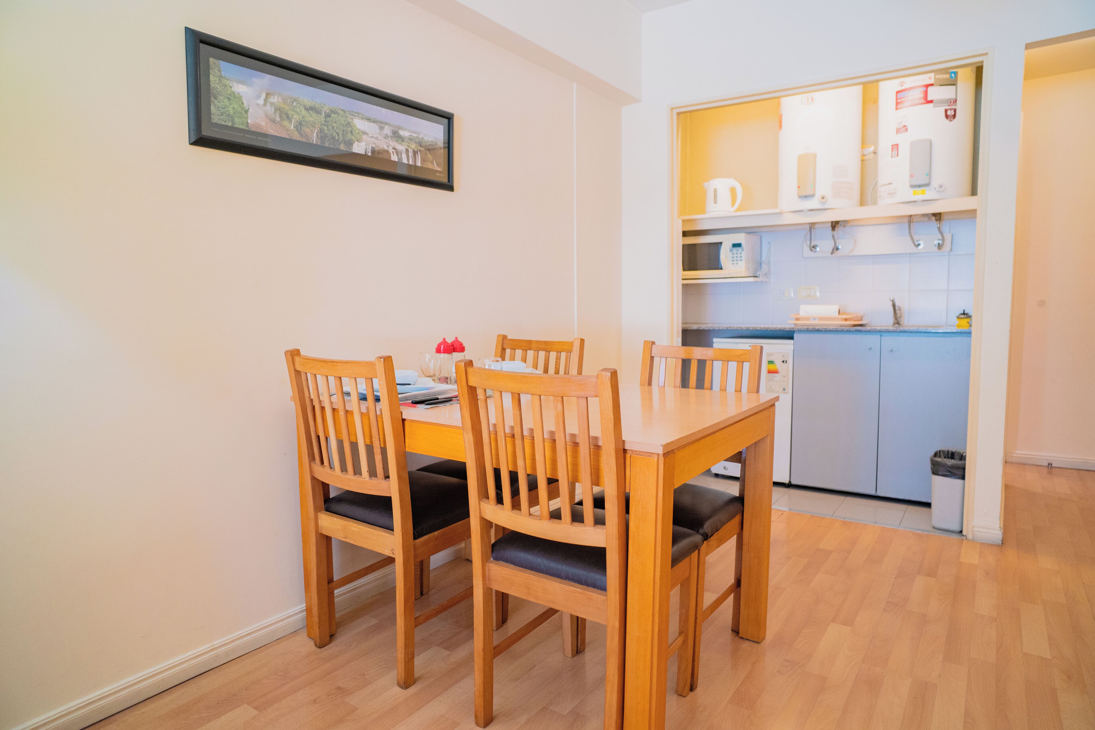 Un comedor con una mesa y sillas de madera, una cocina con armarios y electrodomésticos blancos, y un cuadro en la pared.