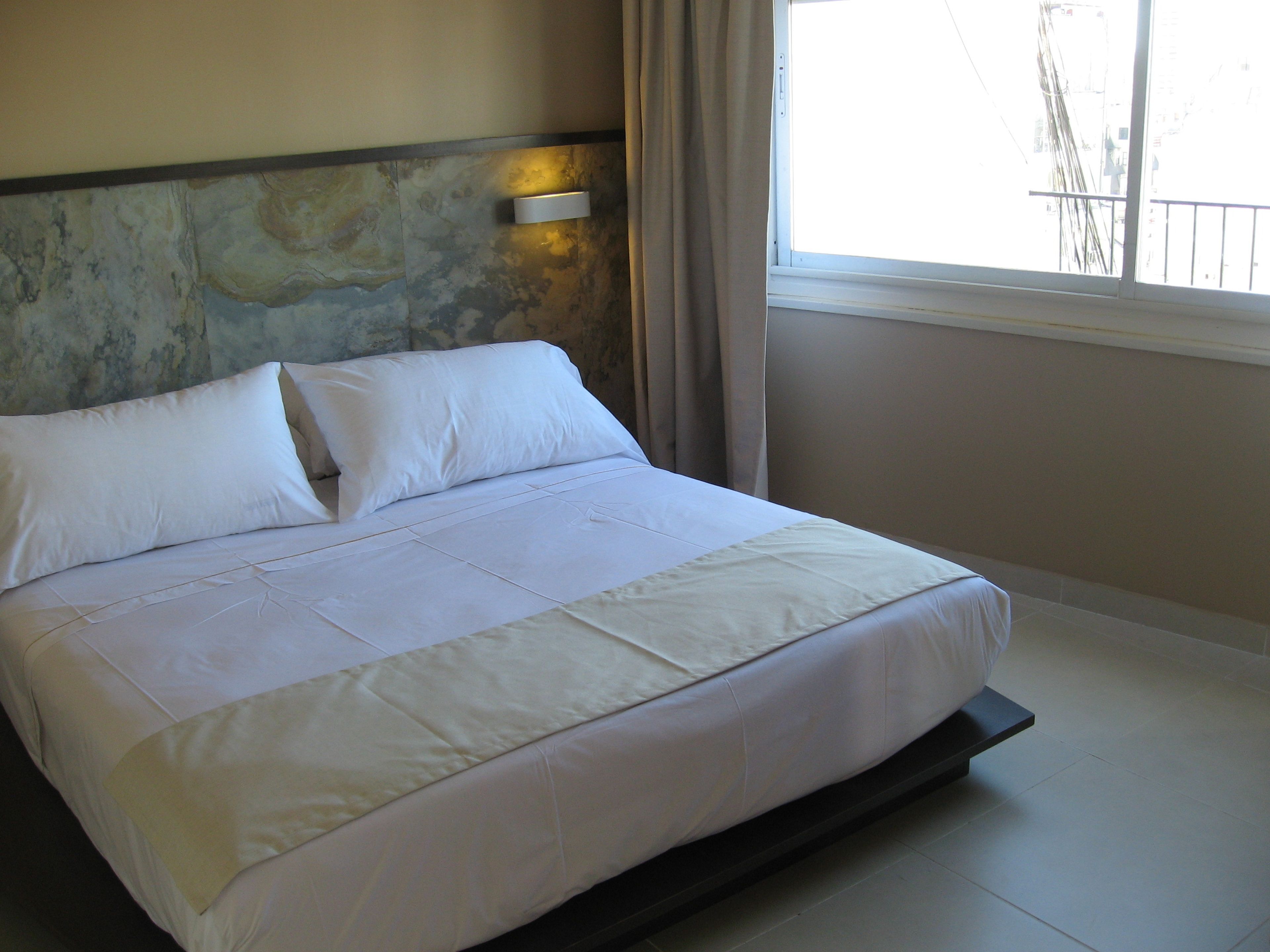 A neatly made bed with white linens, a stone accent wall, a window with curtains, and a view of buildings outside.