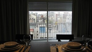 Una mesa de comedor dispuesta con platos, cubiertos y vasos, frente a una ventana con vistas a un paisaje urbano.