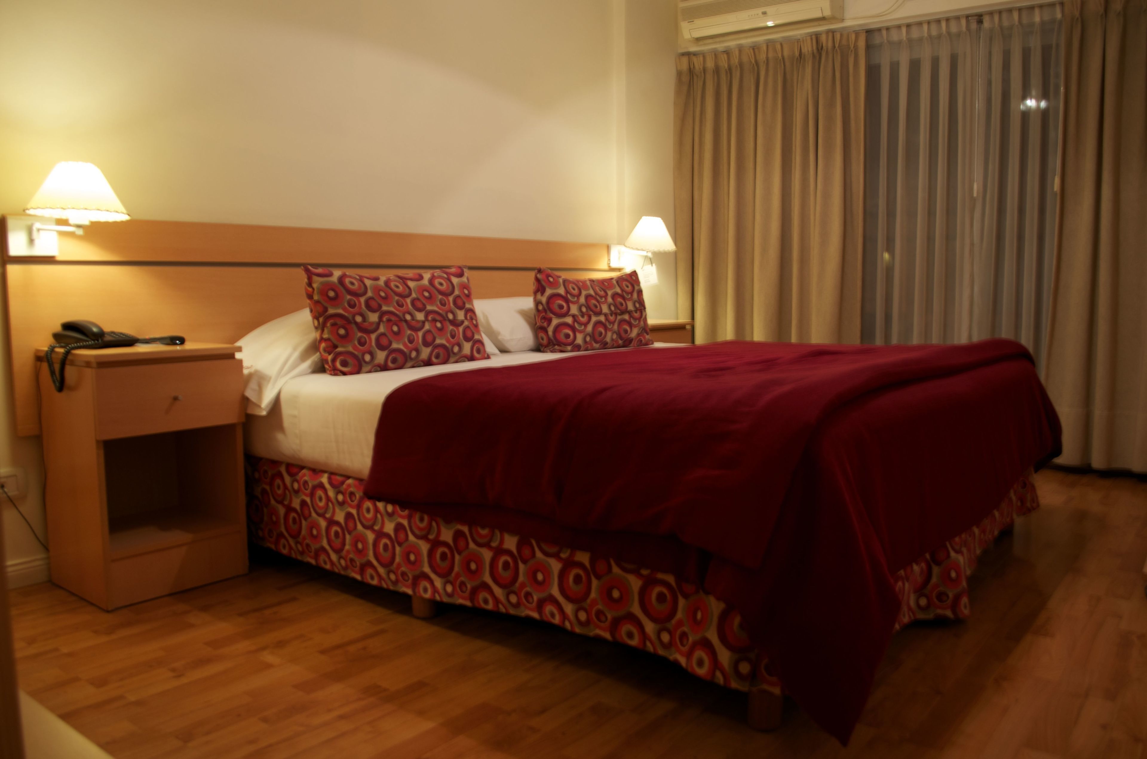 A hotel room with a wooden bed, patterned bedding, two bedside lamps, a nightstand, and a window with curtains.