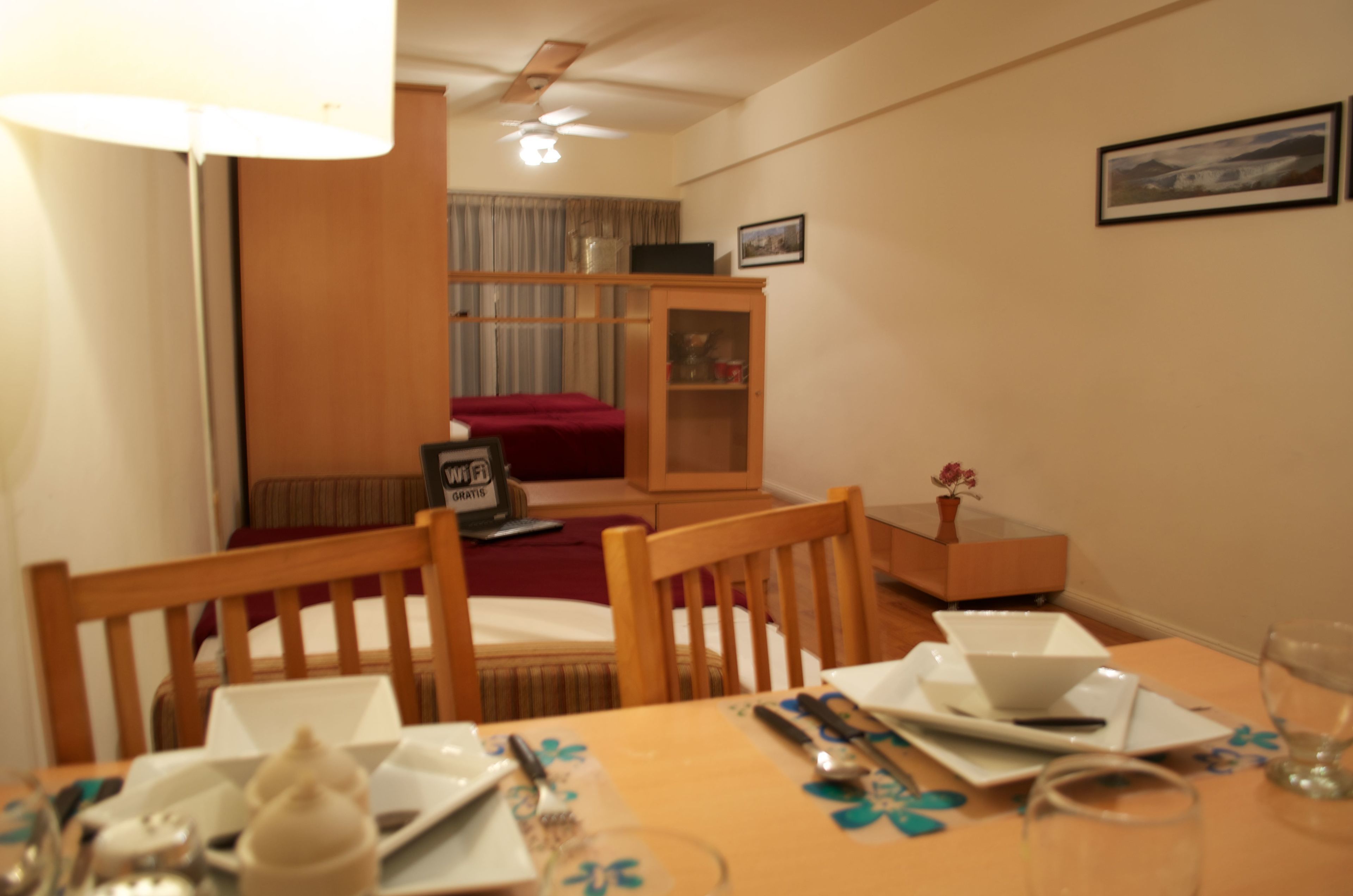 A dining area with a table set for two, a wooden cabinet, and a bed in the background.