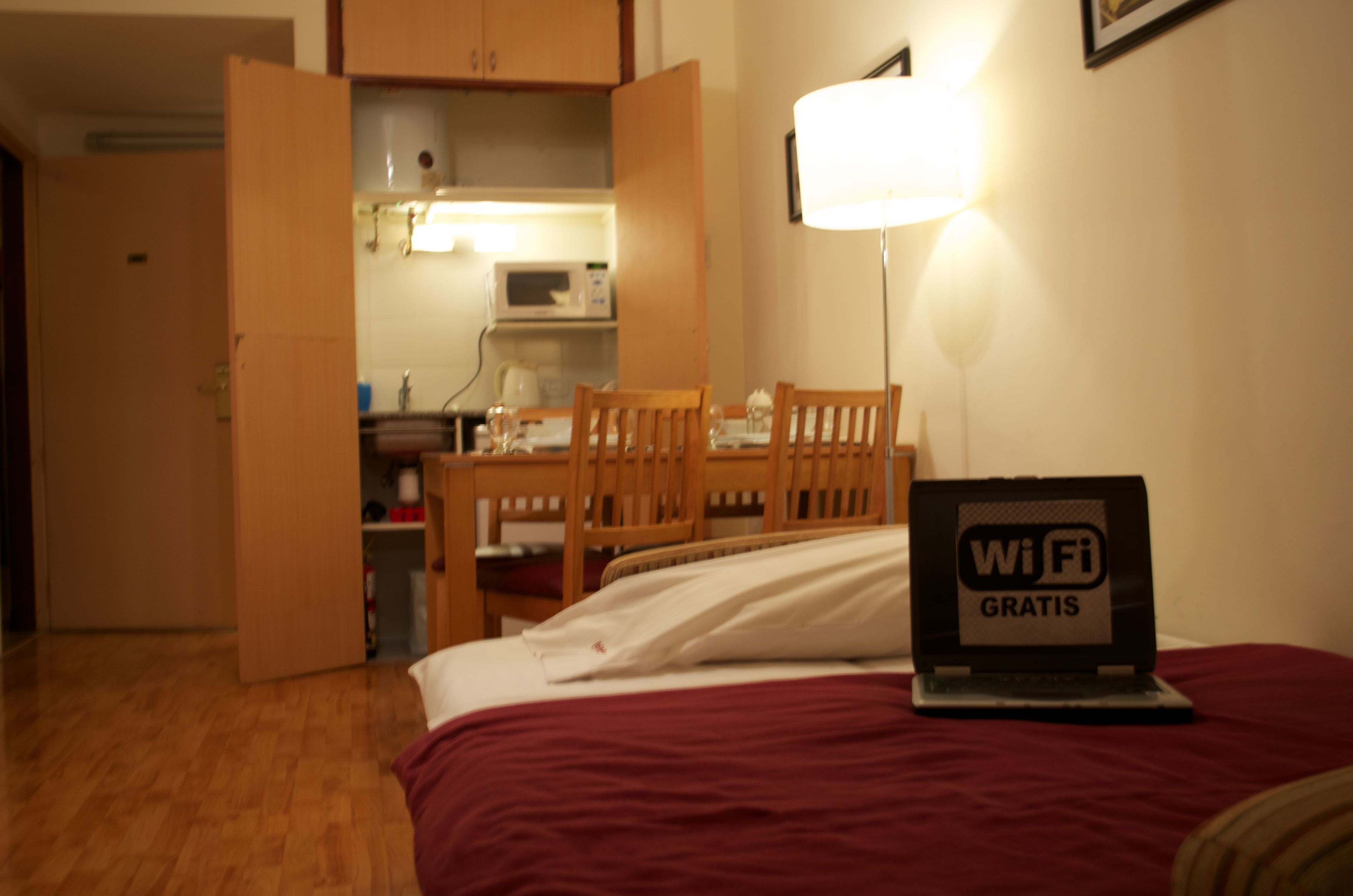 Habitación de hotel con cama, escritorio con una laptop mostrando “WiFi Gratis”, una silla, una mesa y un microondas al fondo.