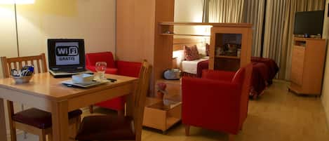 A compact hotel room with a wooden desk, a laptop, a red sofa, a bunk bed, and a TV.