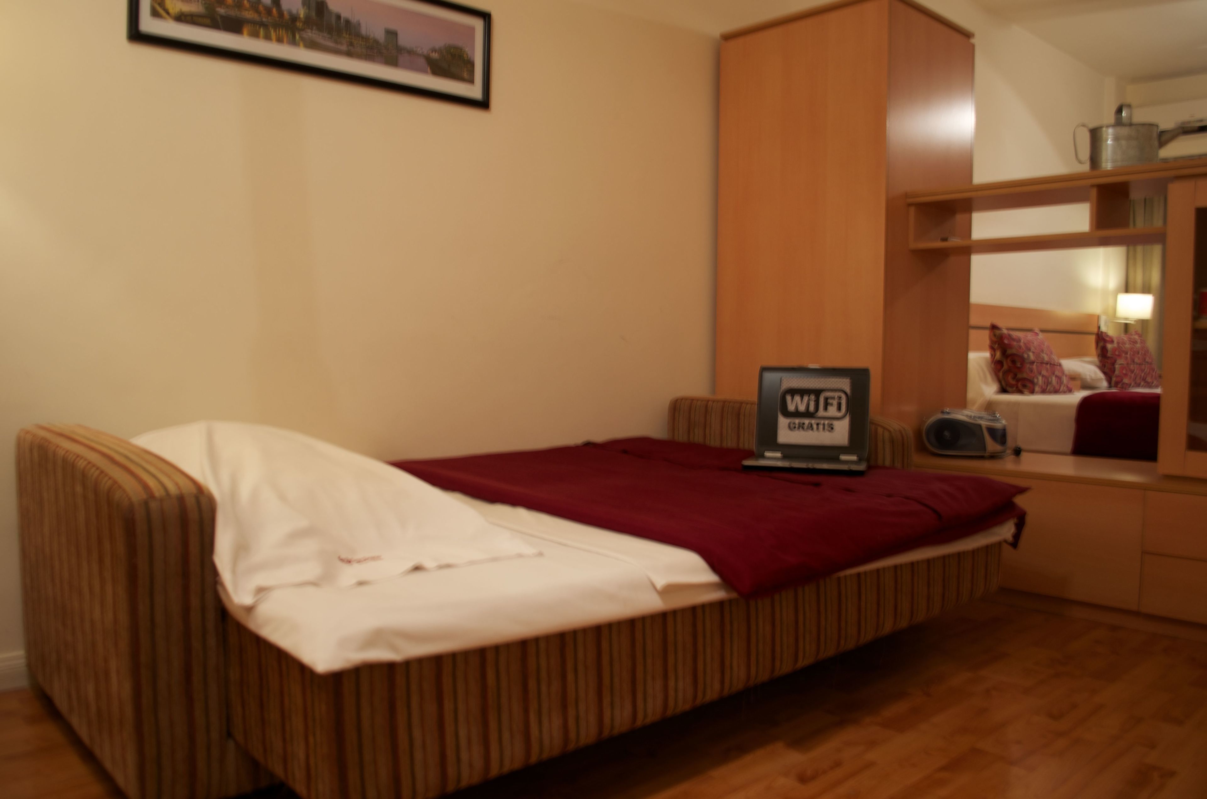 A single bed with a red blanket, a small bedside table with a radio, and a sign indicating Wi-Fi availability.