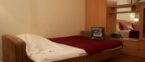 A single bed with a red blanket, a small bedside table with a radio, and a sign indicating Wi-Fi availability.