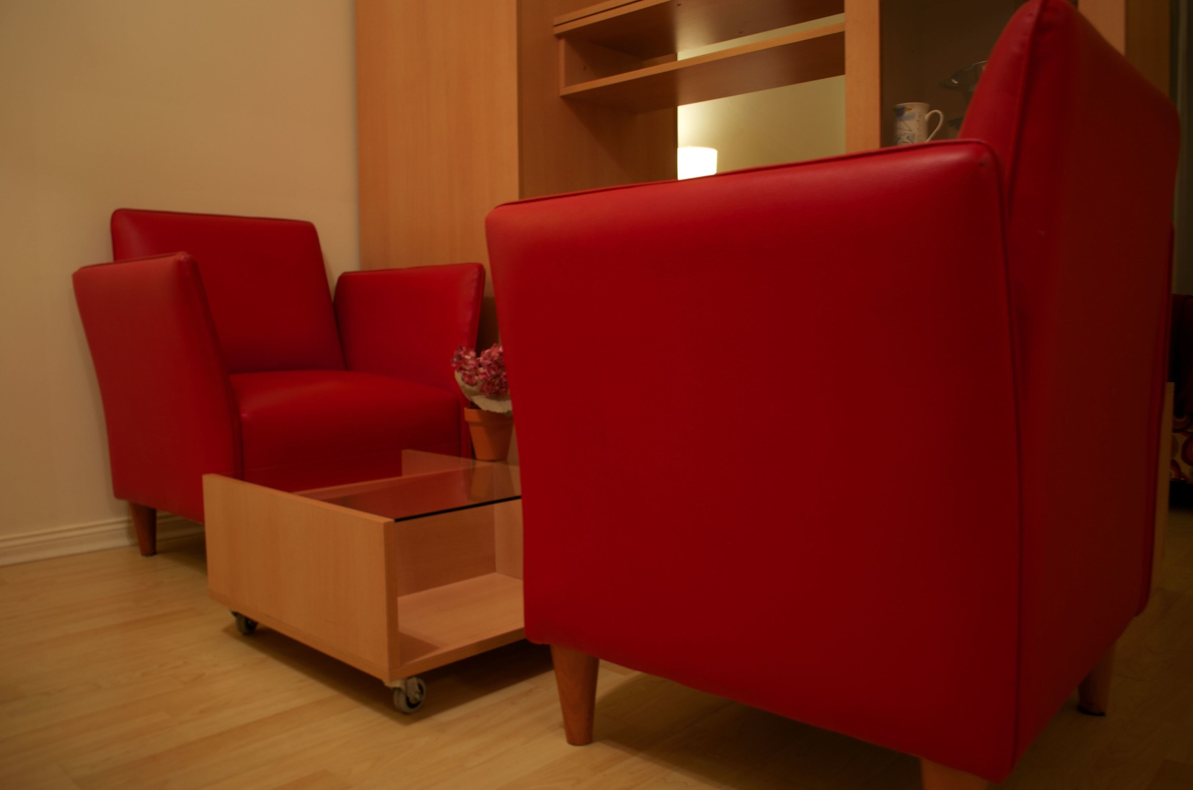 Ein roter Sessel und ein kleiner Glastisch in einem Raum mit Holzboden und einem Holzschrank im Hintergrund.