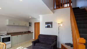 A modern kitchen with white cabinets, a microwave, and a stove. There's a gray sofa and a black chair in the living area, with a wooden staircase leading to another level. - ASURE Chelsea Gateway Motor Lodge (Westport)