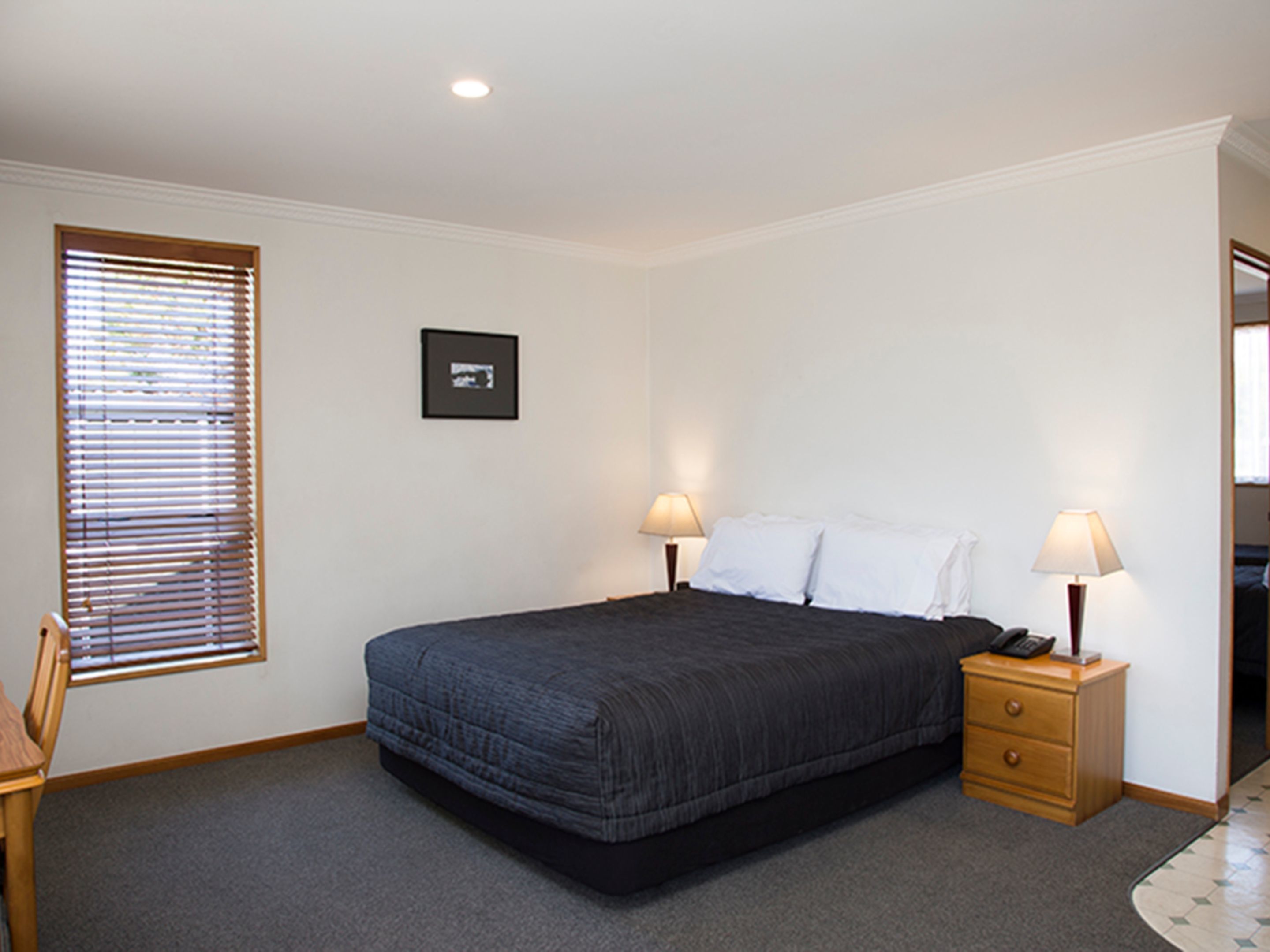 Une chambre à coucher avec un lit, deux tables de chevet avec des lampes, une chaise, un tableau encadré au mur et une fenêtre avec des stores.
