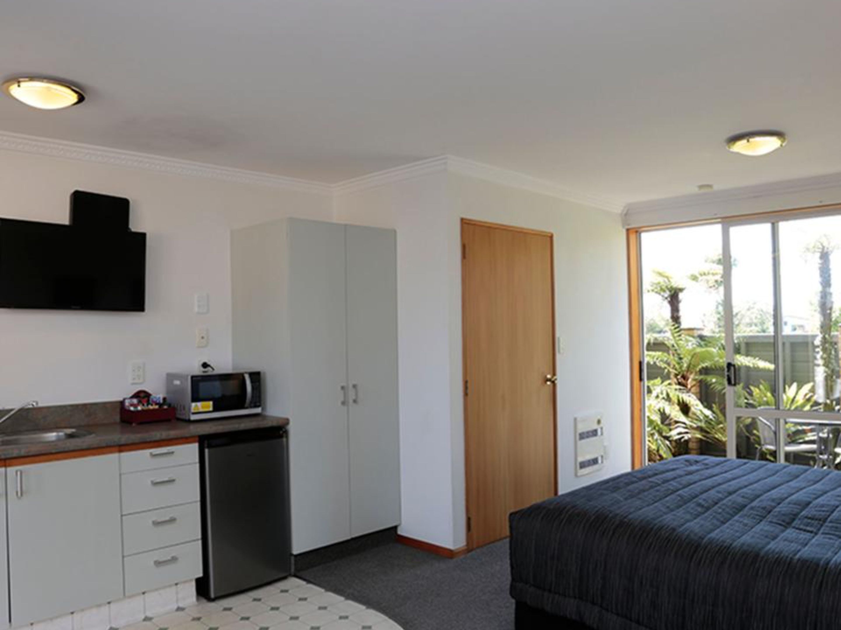 Une chambre avec un lit, une kitchenette équipée d’un micro-ondes et de placards, et une porte donnant sur un balcon agrémenté de plantes.