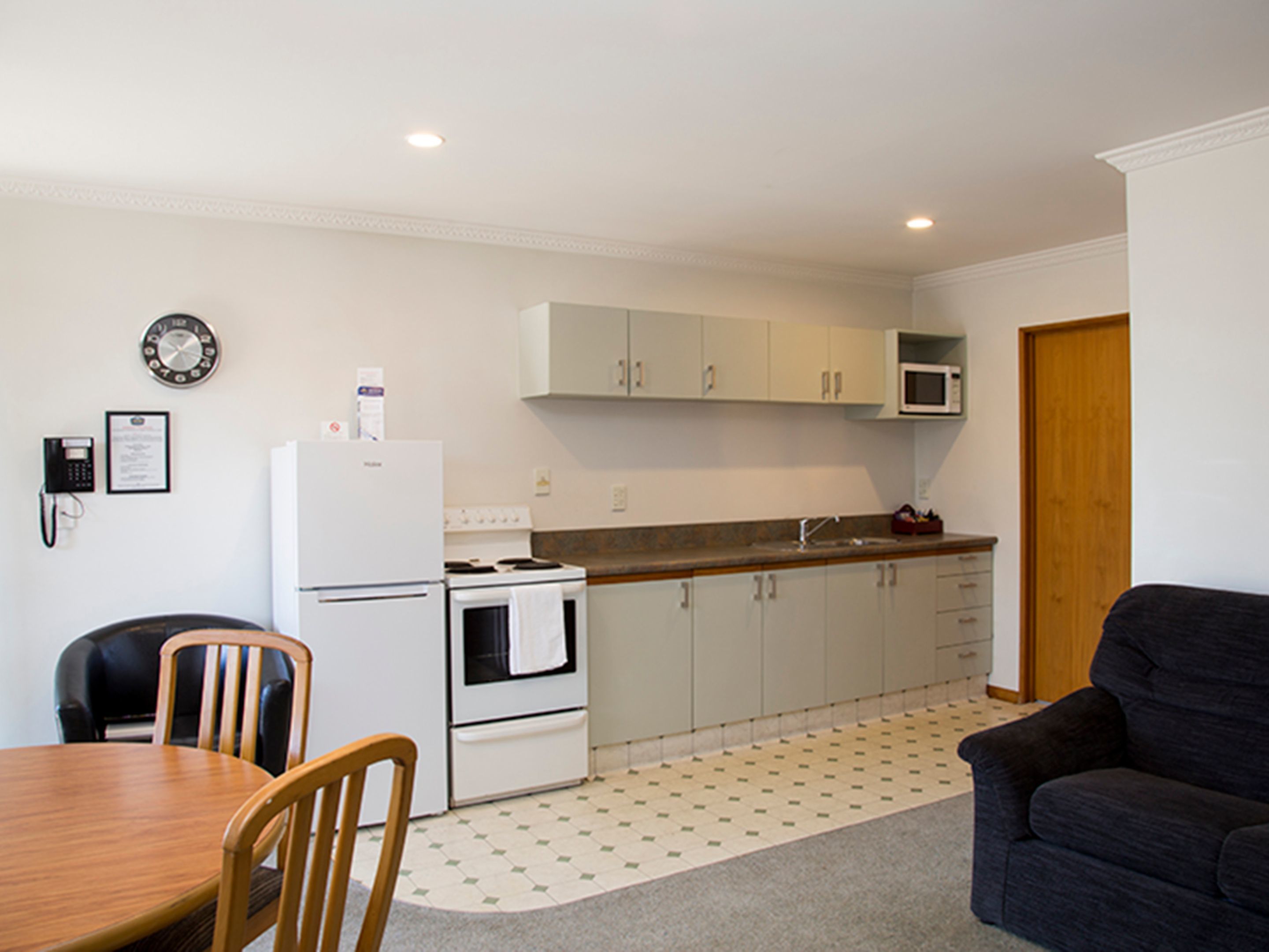 A compact kitchen with white cabinets, a microwave, a refrigerator, and a stove. There is a dining table with chairs and a dark-colored sofa in the living area.
