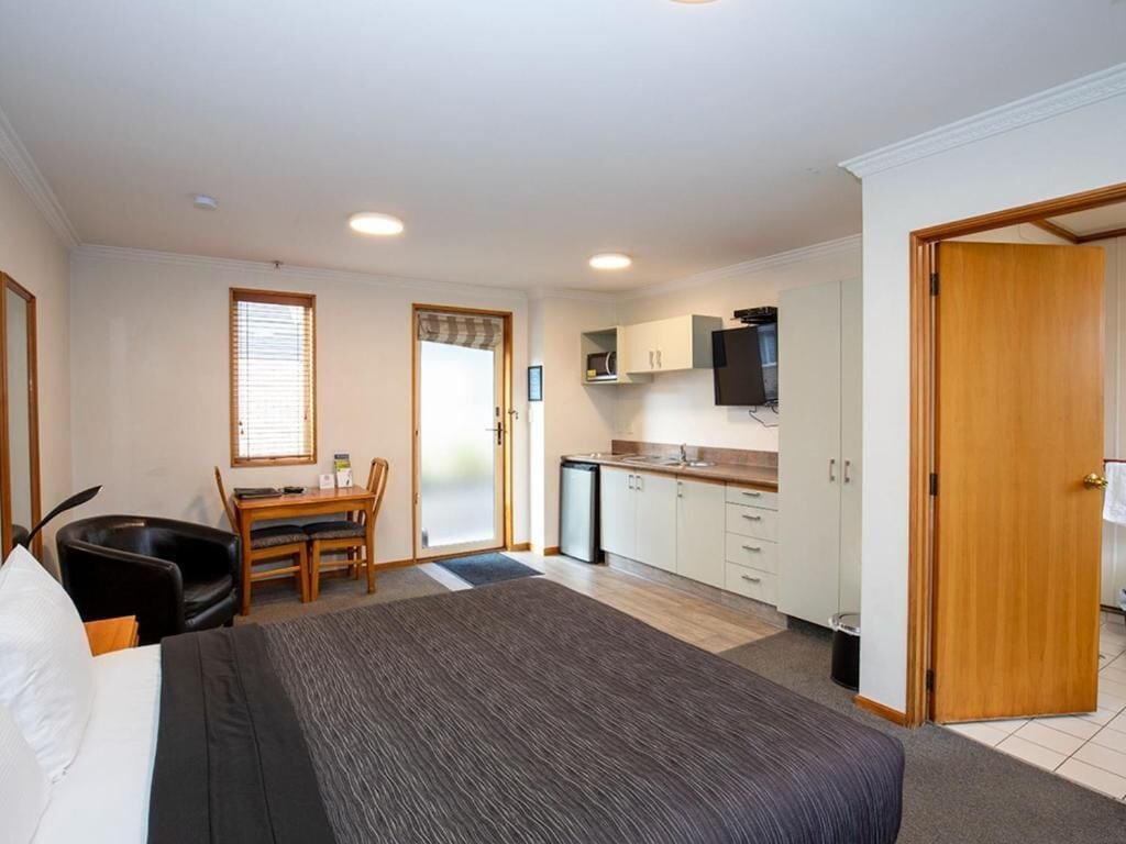 A hotel room with a bed, a desk, a chair, a kitchenette, and a door.