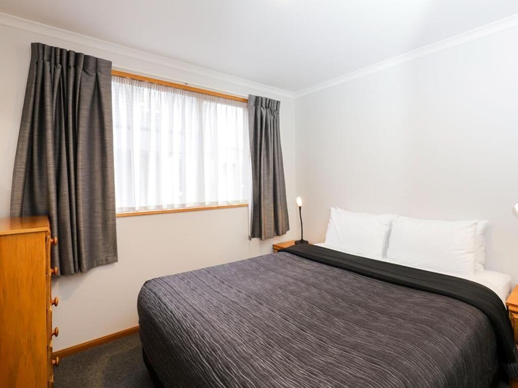 A hotel room with a bed, a wooden nightstand, a lamp, and a window with curtains.