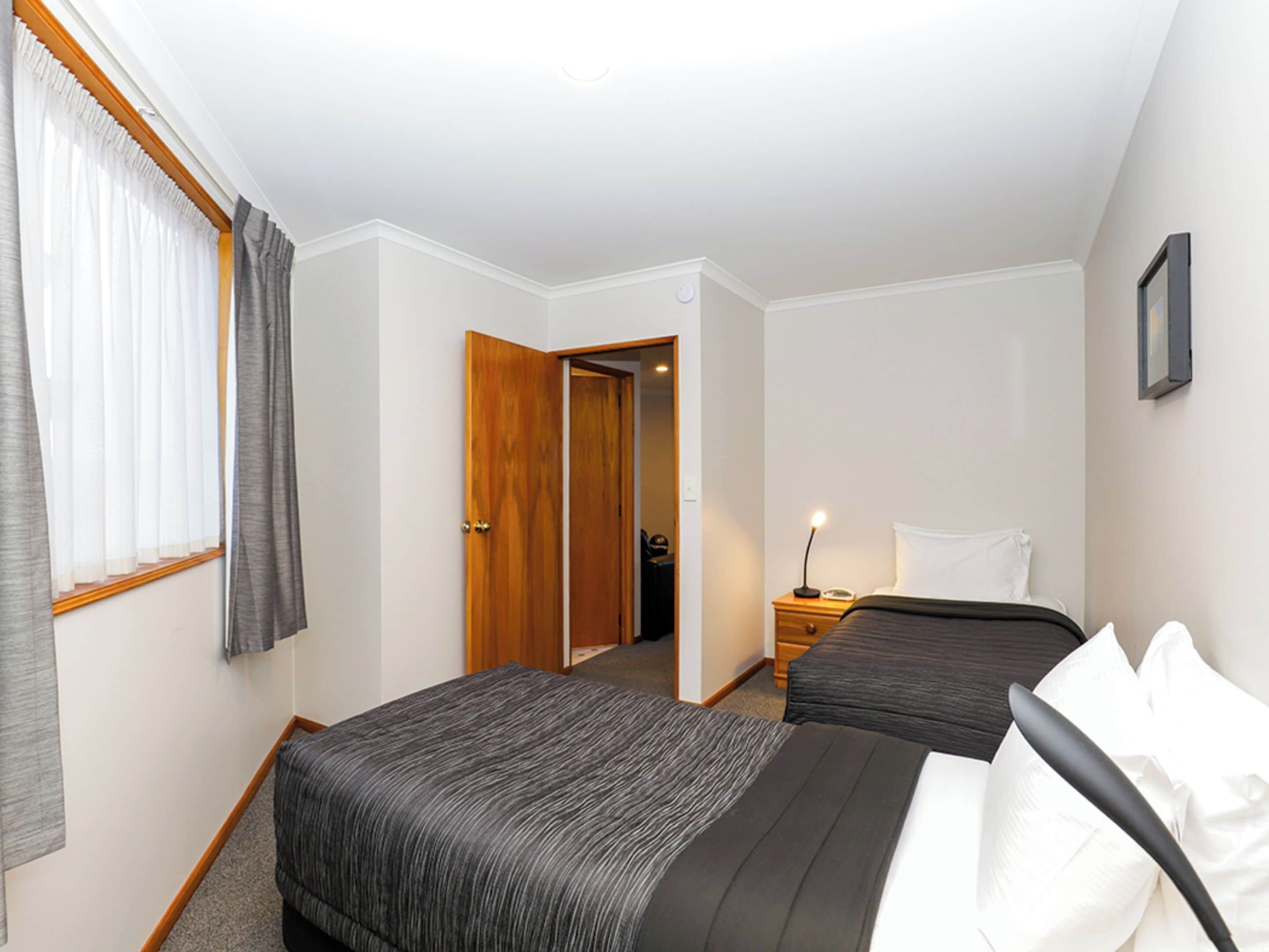 A hotel room with two beds, a nightstand with a lamp, a door leading to another room, and a window with curtains.
