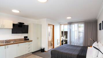 Quarto de hotel moderno com uma cama grande, uma cozinha compacta, uma televisão de tela plana e um banheiro visível por uma porta aberta.