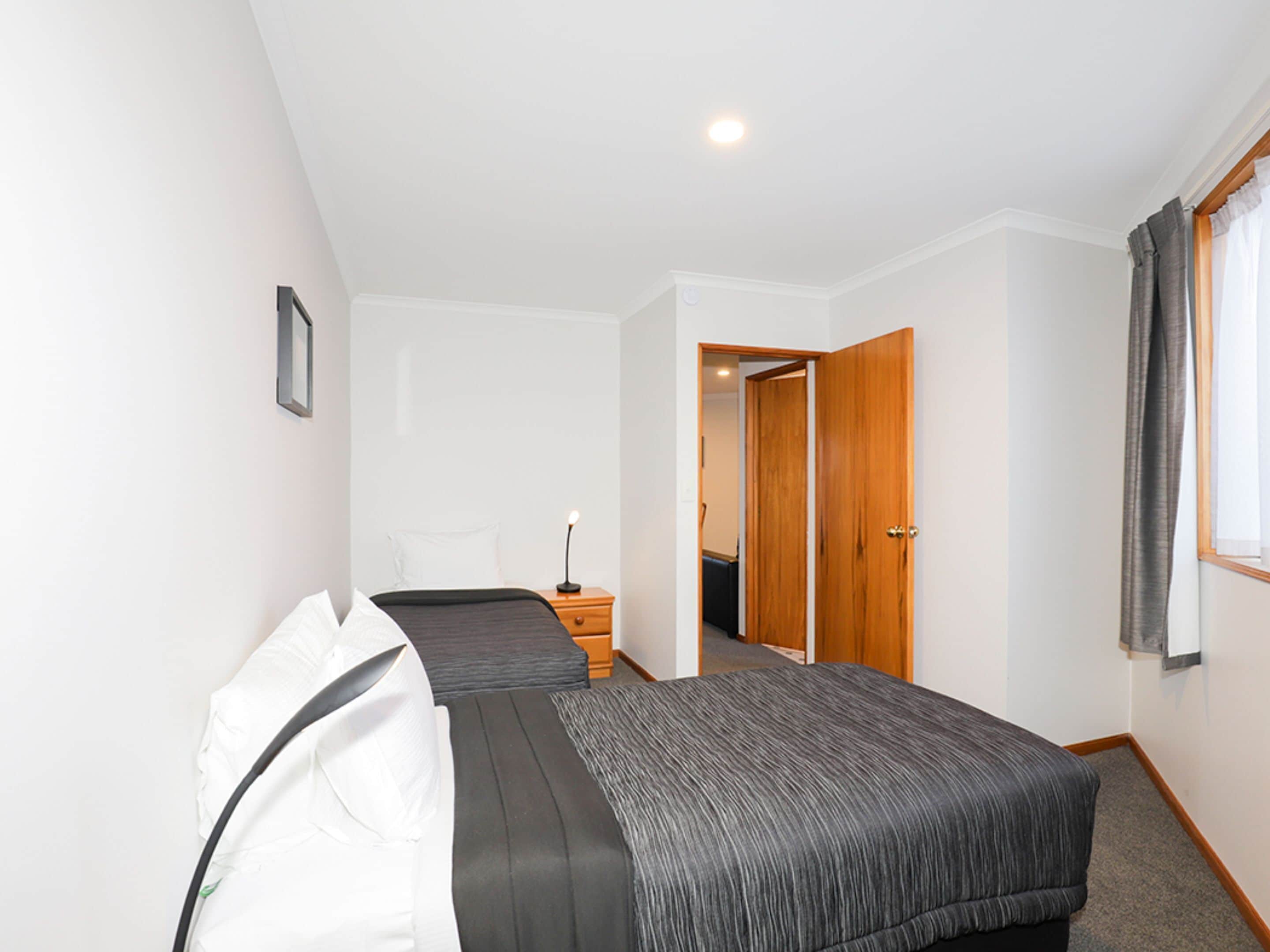 A hotel room with two beds, a bedside table with a lamp, a wooden door, and a window with curtains.