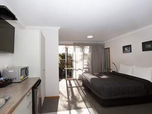 A compact hotel room with a kitchenette, a bed, a TV, and a balcony with a view of greenery. - ASURE Chelsea Gateway Motor Lodge (Westport)