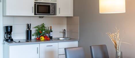 A modern kitchen with white cabinets, a microwave, a coffee maker, and a sink. There is a dining table with chairs and a vase with dried plants.