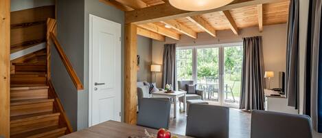 Un comedor con una mesa de madera, sillas y un bol de manzanas. Se ve una escalera con barandillas de madera y una puerta blanca. El cuarto tiene techo de madera y ventanas grandes con cortinas.