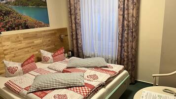 Een keurig opgemaakt bed met geruit beddengoed en kussens met hartjesprint, een houten hoofdbord, uitzicht op een meer door dunne gordijnen en een tafeltje met een menukaart.