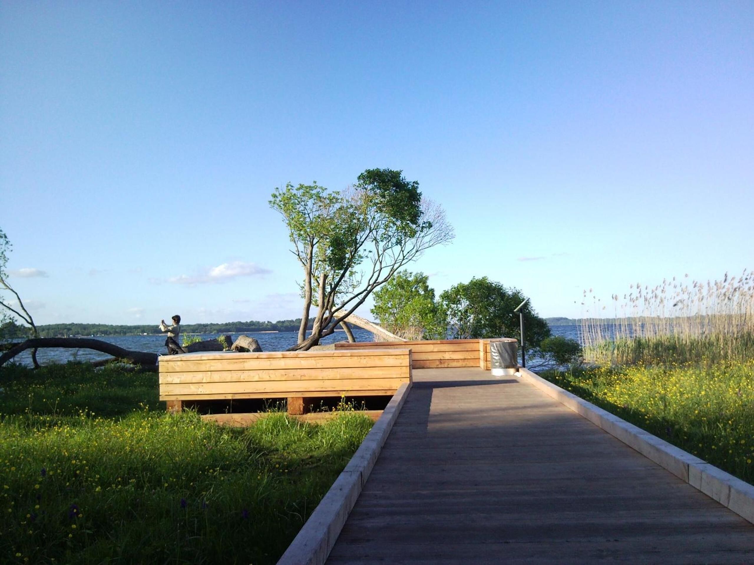 Une terrasse en bois mène à une structure également en bois, dotée de bancs, et offre une vue sur une étendue d’eau.