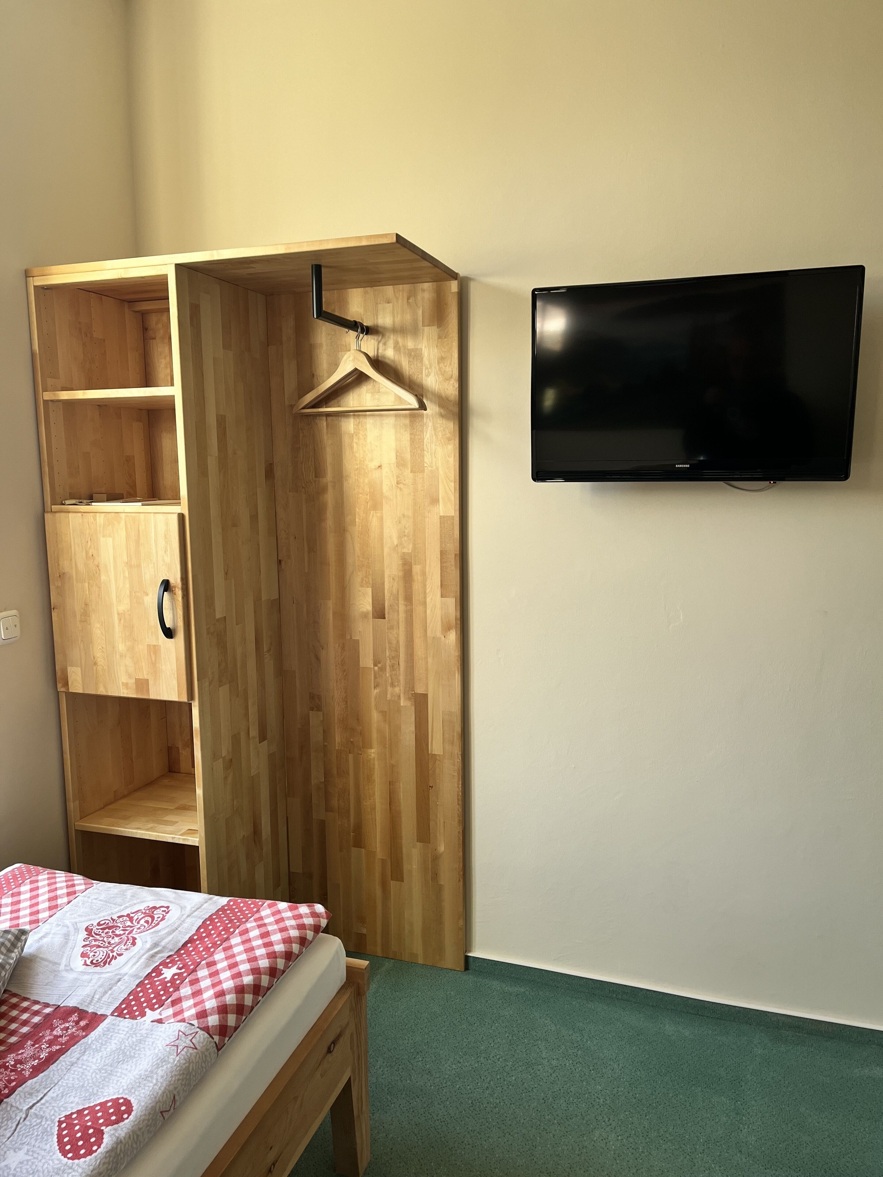 A bedroom with a wooden wardrobe, a bed with a checkered bedspread, and a mounted TV on the wall.