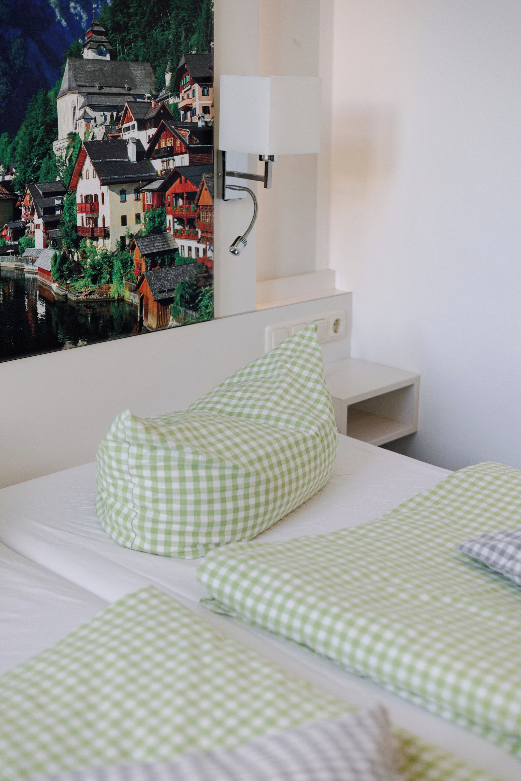 Een slaapkamer met een bed, beddengoed met groene ruitjes, een nachtkastje, een wandlamp en een schilderij van een dorp.