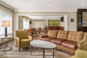 A hotel lobby with a yellow armchair, a brown leather sofa, and a round table. There is a dining area in the background with a green wall and a television.